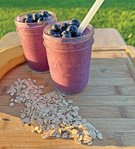 Two blueberry muffin smoothies are displayed on wood cutting board with raw oats and banana.