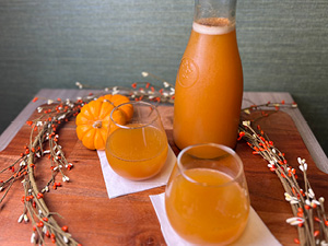 Two glasses of pumpkin spice mocktail next to a full carafe served on a wood cutting board with mini pumpkins.