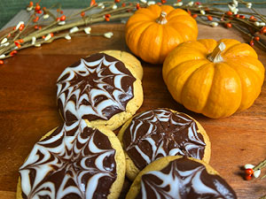 Four spider web cookies are displayed with mini pumpkins on a wood cutting board.