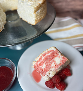 Angel Food Cake with Raspberry Drizzle is displayed on white plate with fresh berries on the side and the full cake in the background.