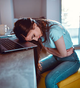 A young woman at work is hunched over in pain resting her head on her laptop.
